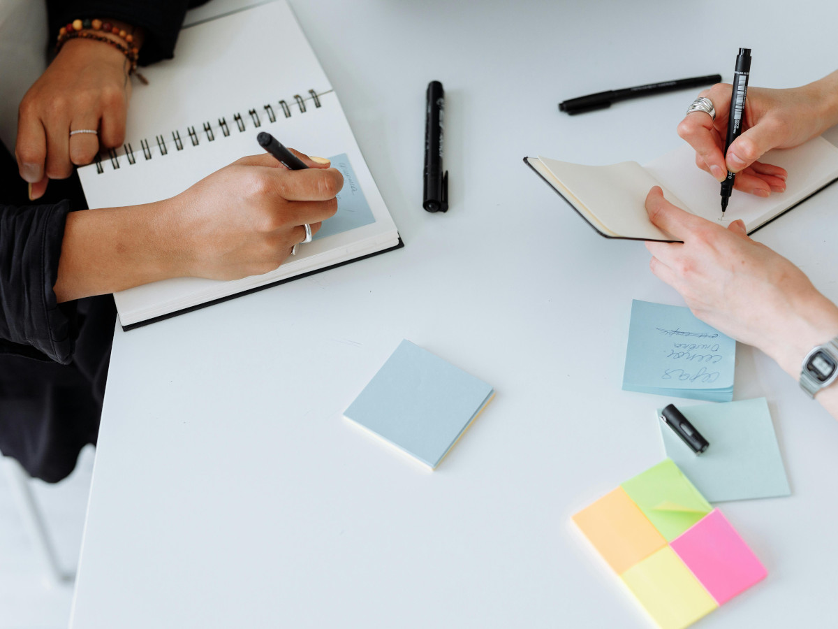 An image of a close up of a desk where there are two sets of hands writing in two notebooks with lots of post its. Photo by Thirdman : https://www.pexels.com/photo/people-writing-on-papers-8485658/